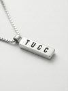 Silver necklace with a bar pendant displaying 'TUCC' on a light gray background