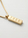 Gold necklace with a bar pendant engraved with 'TUCC' on a light gray background