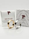 Two rings, one gold and one silver, in front of Tucc packaging on a white background.